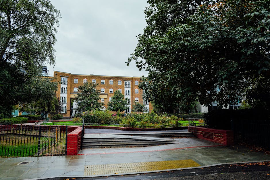 A view of an outdoor staircase leading into a residential area with multi-story brick flats. The staircase is made of concrete with metal handrails and is partially blocked by a black metal fence. The pavement in the foreground is wet, reflecting recent rain, and features a yellow tactile paving strip for accessibility. Surrounding the staircase, there are green trees and well-maintained garden beds with flowering plants, providing a lush environment. Natural light from an overcast sky illuminates the scene, highlighting the clean and orderly appearance of the area. This image is associated with Harringay Carpet Cleaners, illustrating the importance of surface cleaning and maintenance even in outdoor communal spaces, fitting within the context of access issues in Harringay flats and carpet cleaning solutions.
