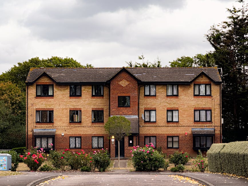 Image of a three-story brick residential building in Harringay with a symmetrical facade featuring six windows on each floor, some with black window frames and white curtains, set against a backdrop of trees and a cloudy sky. The entrance door is centrally located, with a small tree and well-maintained pink flowering shrubs in front. The surrounding pavement and driveway are clean, and the overall appearance suggests good exterior maintenance and hygiene. This setting relates to the topic of access issues and cleaning solutions for Harringay flats, emphasizing the importance of surface cleaning and upkeep by Harringay Carpet Cleaners.