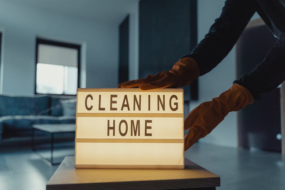 A person wearing brown cleaning gloves is adjusting a lightbox with the words 'CLEANING HOME' illuminated in black lettering on white background panels. The scene takes place in a modern living room with a grey sofa, coffee table, and large windows that allow natural light to fill the space. The surface of the lightbox is a smooth, flat surface, and the room appears clean and tidy, emphasizing hygiene and maintenance suitable for domestic cleaning services. Harringay Carpet Cleaners' branding and the context of avoiding hidden cleaning charges are subtly implied through the setup, highlighting professional surface cleaning and deep cleaning processes in residential environments.