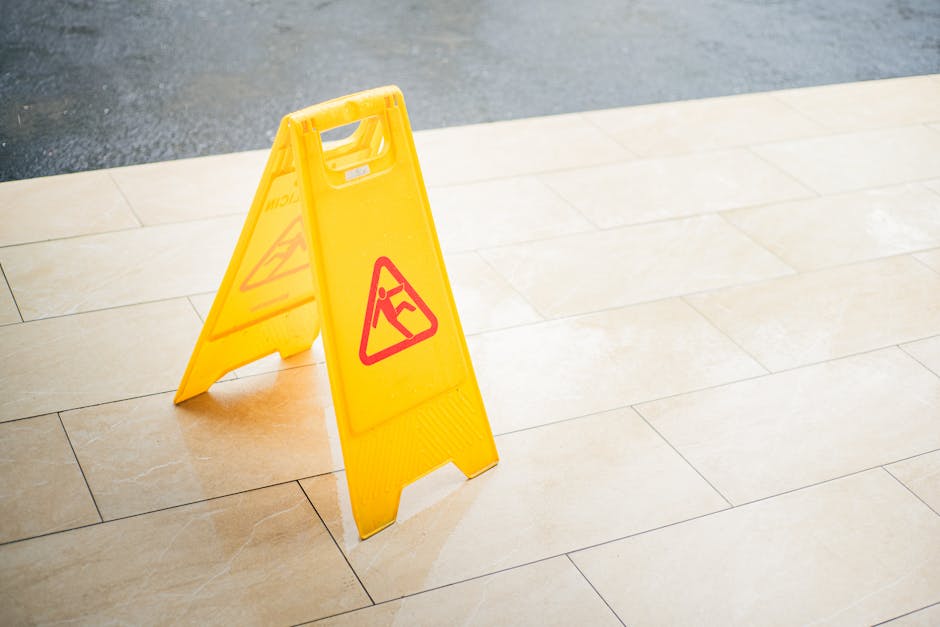 A bright yellow portable wet floor caution sign with a red warning symbol and text positioned on a clean, light-colored ceramic tiled floor in an indoor or outdoor area. The tiles have a smooth, shiny surface, and the lighting highlights their glossy finish. The sign is placed in the center of the image, indicating that the flooring may be wet or slippery, emphasizing safety during surface cleaning or maintenance. The background shows a portion of darker flooring or pavement, creating contrast with the clean tiles. The setting suggests a commercial or public space, and the image relates to hygienic surface cleaning and safety procedures, aligning with Harringay Carpet Cleaners' services.