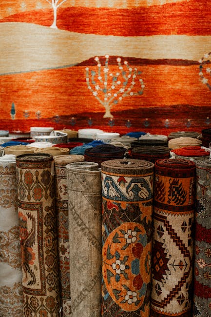 A display of rolled-up traditional woven rugs with intricate geometric and floral patterns in various colors such as beige, brown, red, and blue, arranged upright on a surface in front of a large, colorful tapestry with a stylized tree design and abstract landscape. The setting suggests an indoor environment, possibly a shop or showroom, where surface cleaning and maintenance of textiles is emphasized. The rugs appear clean and well-maintained, reflecting professional cleaning standards offered by Harringay Carpet Cleaners, as indicated on the webpage about Finsbury Park rug cleaning and collection in Harringay.