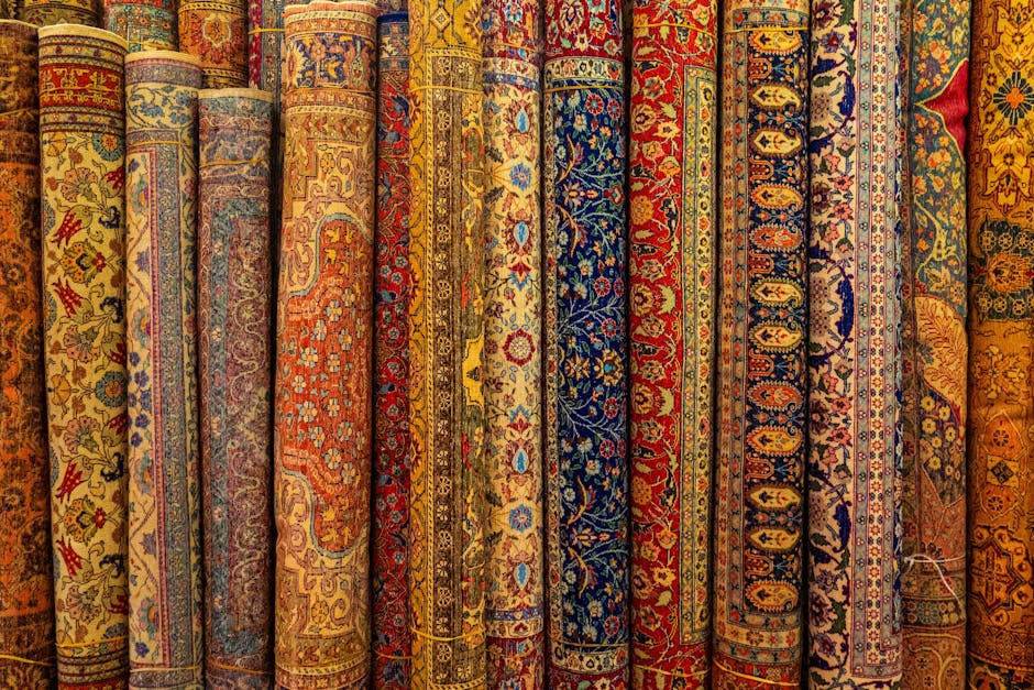 A display of rolled-up traditional woven rugs with intricate geometric and floral patterns in various colors such as beige, brown, red, and blue, arranged upright on a surface in front of a large, colorful tapestry with a stylized tree design and abstract landscape. The setting suggests an indoor environment, possibly a shop or showroom, where surface cleaning and maintenance of textiles is emphasized. The rugs appear clean and well-maintained, reflecting professional cleaning standards offered by Harringay Carpet Cleaners, as indicated on the webpage about Finsbury Park rug cleaning and collection in Harringay.
