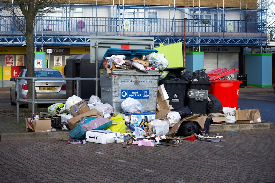 An overflowing large grey mixed recycling bin filled with various papers, cardboard, and plastic waste sits on a sidewalk, surrounded by additional black and red rubbish bags and scattered recyclable 
