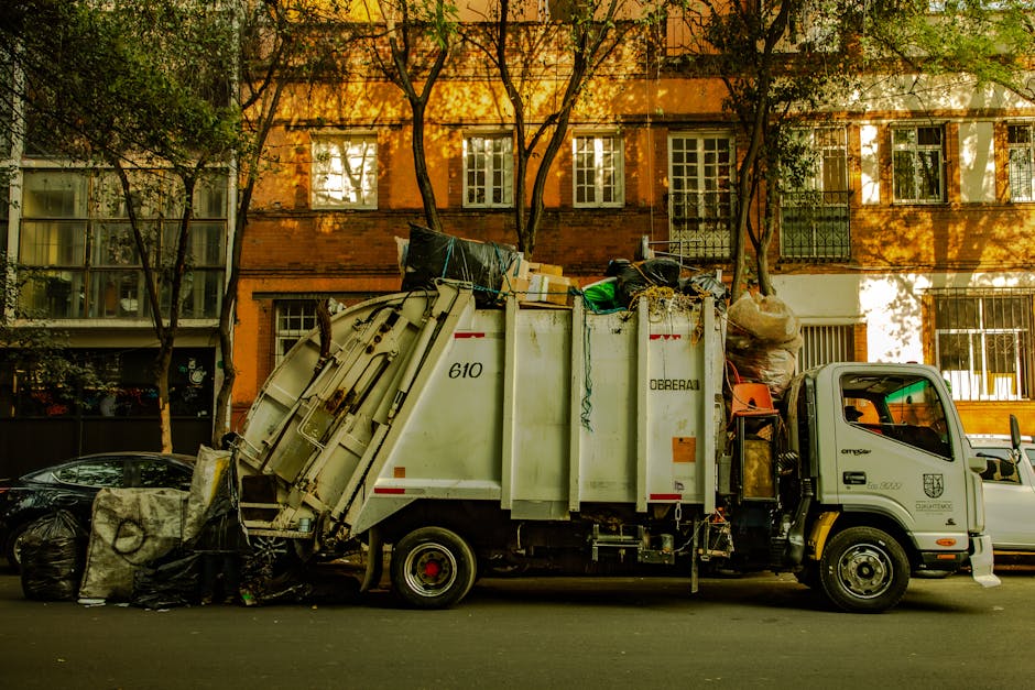 An overflowing large grey mixed recycling bin filled with various papers, cardboard, and plastic waste sits on a sidewalk, surrounded by additional black and red rubbish bags and scattered recyclable 