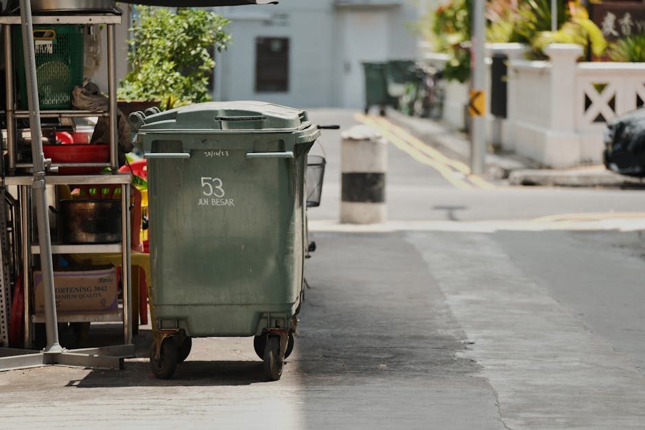 A green wheeled trash bin with the number 53 and the words 'JUN BESAR' printed on the front, positioned on a sidewalk near a storage rack filled with cleaning supplies and tools. The concrete pavement