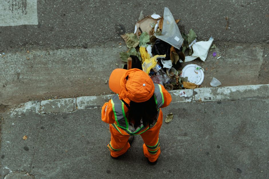 A green wheeled trash bin with the number 53 and the words 'JUN BESAR' printed on the front, positioned on a sidewalk near a storage rack filled with cleaning supplies and tools. The concrete pavement