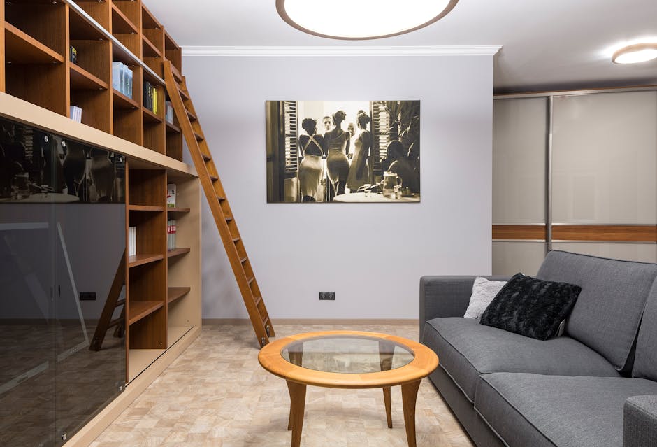A modern living room featuring a grey fabric sofa with a black textured cushion and a white cushion, positioned next to a light wooden coffee table with a glass top. To the left, a large wooden bookshelf with multiple compartments holds books and decorative items, and a wooden ladder is leaned against it for accessing upper shelves. The room has light-colored hardwood flooring and white walls, with a black-and-white framed photograph of women hanging on the wall. On the right side, there are grey sliding doors with wooden trim, and the ceiling has circular recessed lighting. The space is clean, well-maintained, and illuminated by soft natural and artificial light, reflecting professional domestic cleaning and surface sanitisation as part of Harringay Carpet Cleaners' upholstery cleaning and stain removal services.