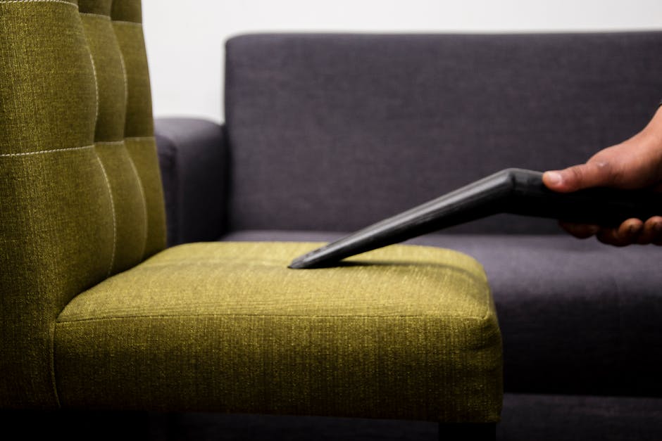 A close-up view of a green upholstered armchair with button-tufted backrest and seat cushion, exhibiting a textured fabric surface. A person is using a handheld vacuum cleaner to perform surface cleaning, targeting the fabric to remove dust and debris. In the background, there is a dark grey sofa made of smooth fabric, positioned against a plain white wall, under bright, even lighting that highlights the cleanliness of the furniture. The scene captures the process of deep cleaning and maintaining hygiene in a residential or commercial setting, with Harringay Carpet Cleaners providing professional upholstery cleaning and stain removal services.