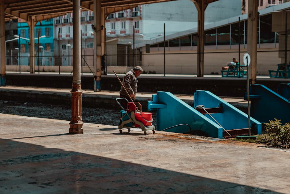A man standing on a concrete platform at Harringay Station, holding a broom and pushing a red cart with cleaning supplies. The platform is partially shaded with sunlight casting shadows, and features blue stairs leading down to the train tracks. In the background, there are benches with a passenger sitting and looking at their phone, modern buildings with glass windows, and a metal fence separating the platform from the tracks. The scene depicts a quiet, well-maintained railway station with clean surfaces and clear signage, representing the type of surface and station cleaning services offered by Harringay Carpet Cleaners. The image highlights the station's urban environment with structural elements such as wooden and metal supports, and the overall cleanliness of the station environment.