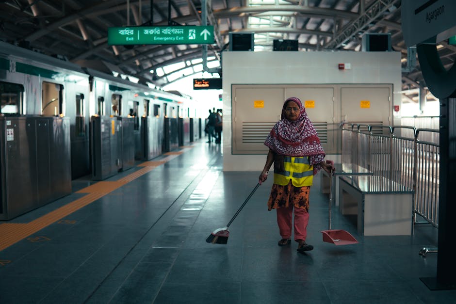 A woman wearing a colorful headscarf, yellow safety vest, and patterned clothing is sweeping the tiled floor of a modern train station platform with a dustpan and broom. The platform features metallic turnstiles, glass barriers, and green signage indicating exit routes and emergency information. Bright natural light illuminates the area, highlighting the clean and well-maintained surfaces. Harringay Carpet Cleaners offers surface cleaning and deep cleaning services, maintaining hygiene and cleanliness in domestic and commercial spaces, as seen in this station cleaning scene.
