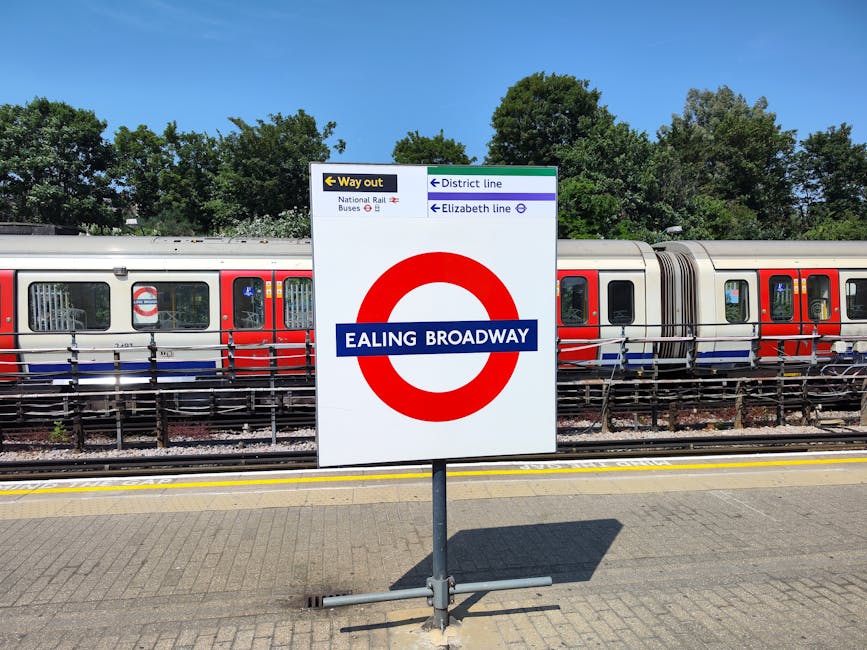 A man standing on a concrete platform at Harringay Station, holding a broom and pushing a red cart with cleaning supplies. The platform is partially shaded with sunlight casting shadows, and features blue stairs leading down to the train tracks. In the background, there are benches with a passenger sitting and looking at their phone, modern buildings with glass windows, and a metal fence separating the platform from the tracks. The scene depicts a quiet, well-maintained railway station with clean surfaces and clear signage, representing the type of surface and station cleaning services offered by Harringay Carpet Cleaners. The image highlights the station's urban environment with structural elements such as wooden and metal supports, and the overall cleanliness of the station environment.
