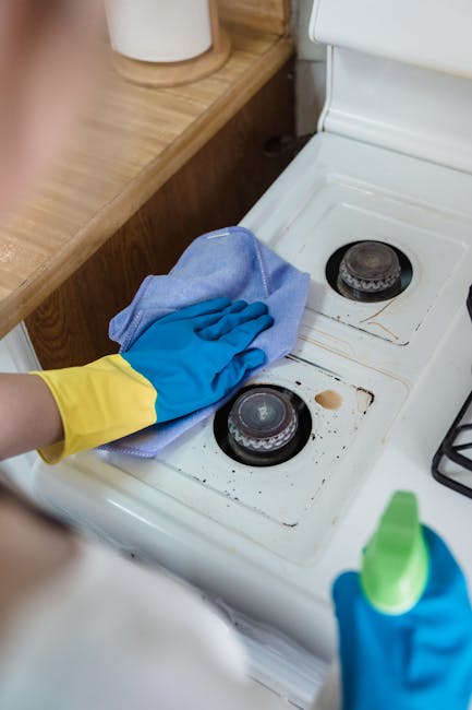 Close-up of a hand using a spray bottle to apply cleaning solution onto a stovetop burner in a kitchen. The stove has two gas burners, one of which is rusty and shows signs of grime, while the other a